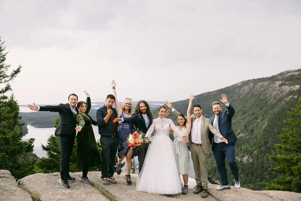 Group of wedding guests posing together on a mountain overlook with forest and water views in the background.