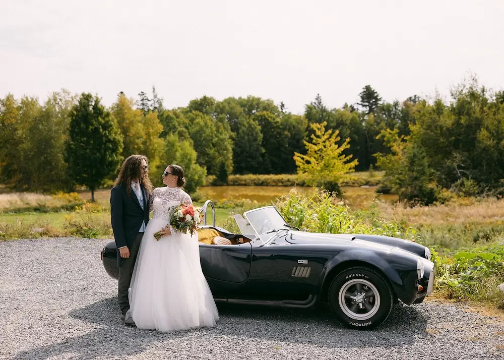 Bride and groom standing together beside a classic, black convertible car in a scenic outdoor setting.