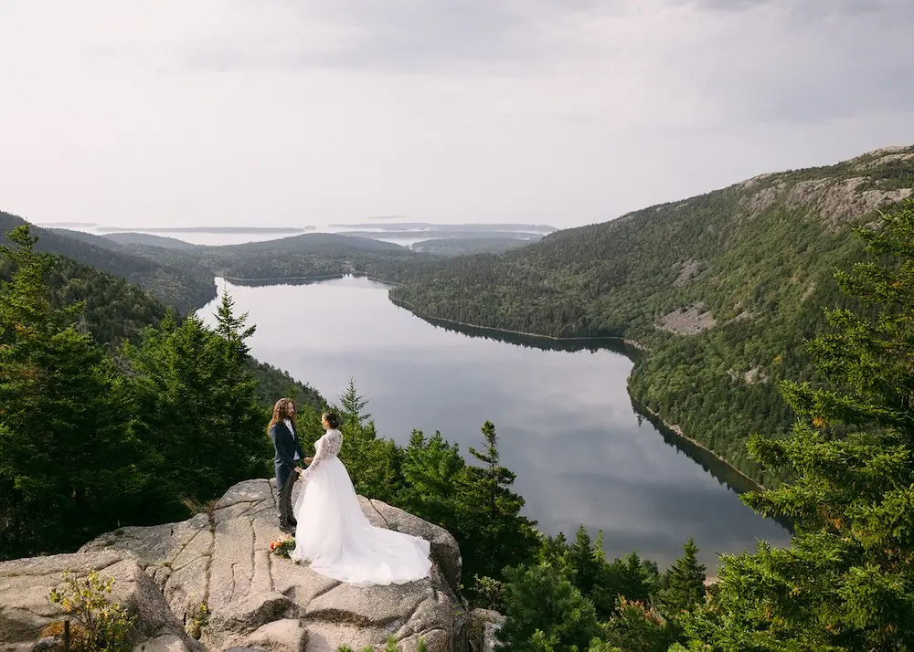 Bride and groom standing on a rocky overlook with a lake and forested mountains in the background.