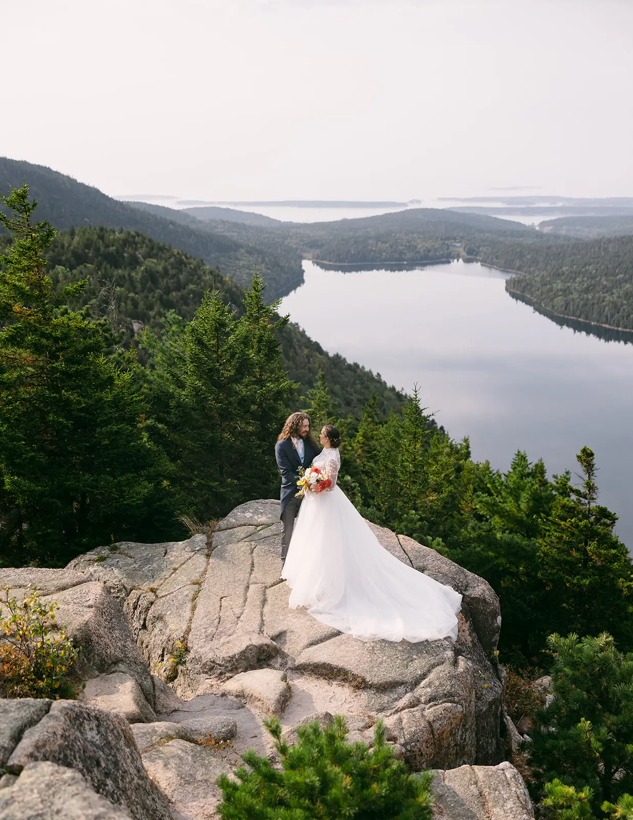 Bride and groom standing on a rocky overlook above a lake at sunrise surrounded by forested mountains.