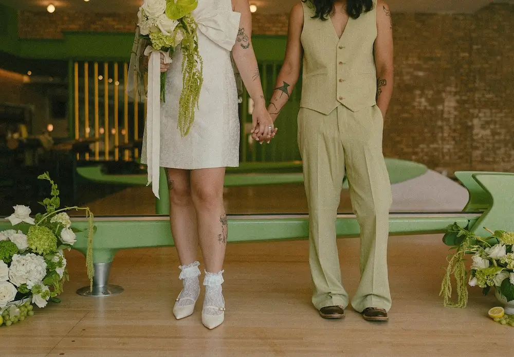 Close-up of a couple holding hands in modern wedding attire at a vintage bowling alley, featuring a short white dress, sage suit, and green-and-white floral arrangements.