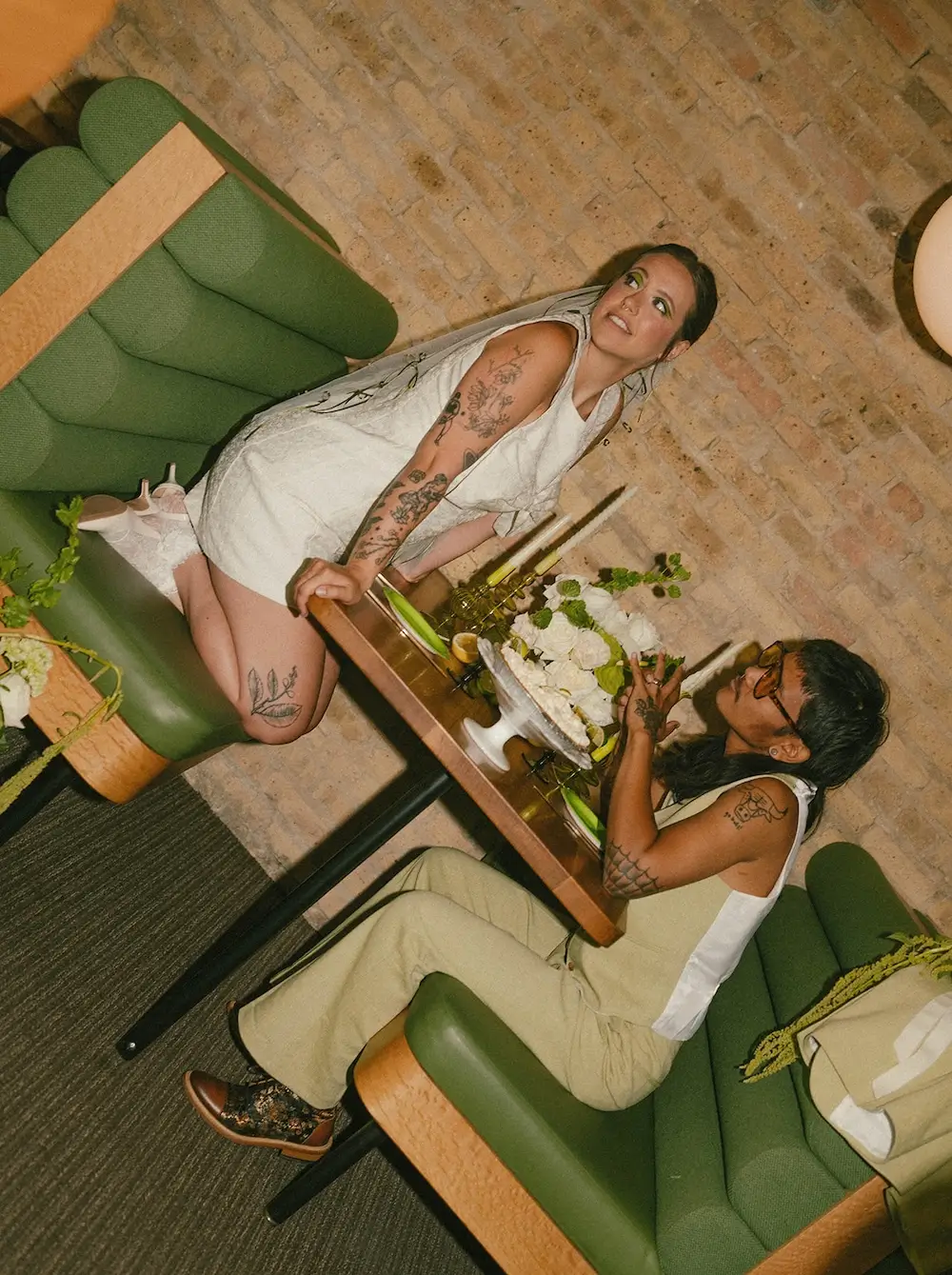 Couple in modern wedding attire seated in a vintage green booth at a mid-century bowling alley, sharing dessert with green and white floral accents on the table.