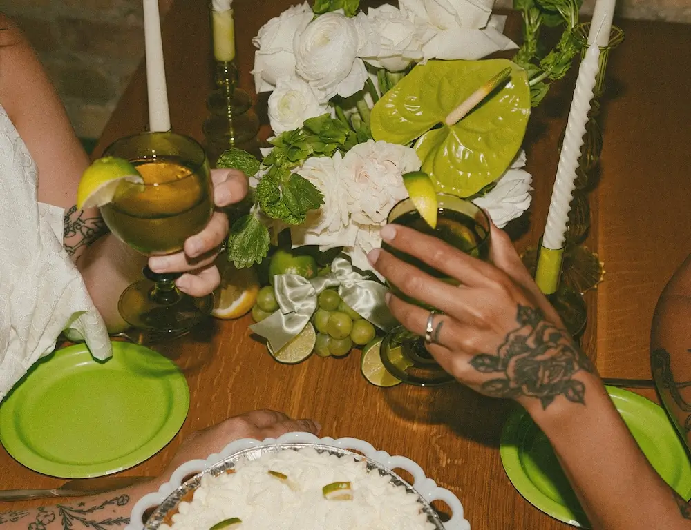 Close-up of a couple toasting with green cocktails at a mid-century bowling alley wedding, surrounded by white florals, citrus slices, and lime-green table accents.