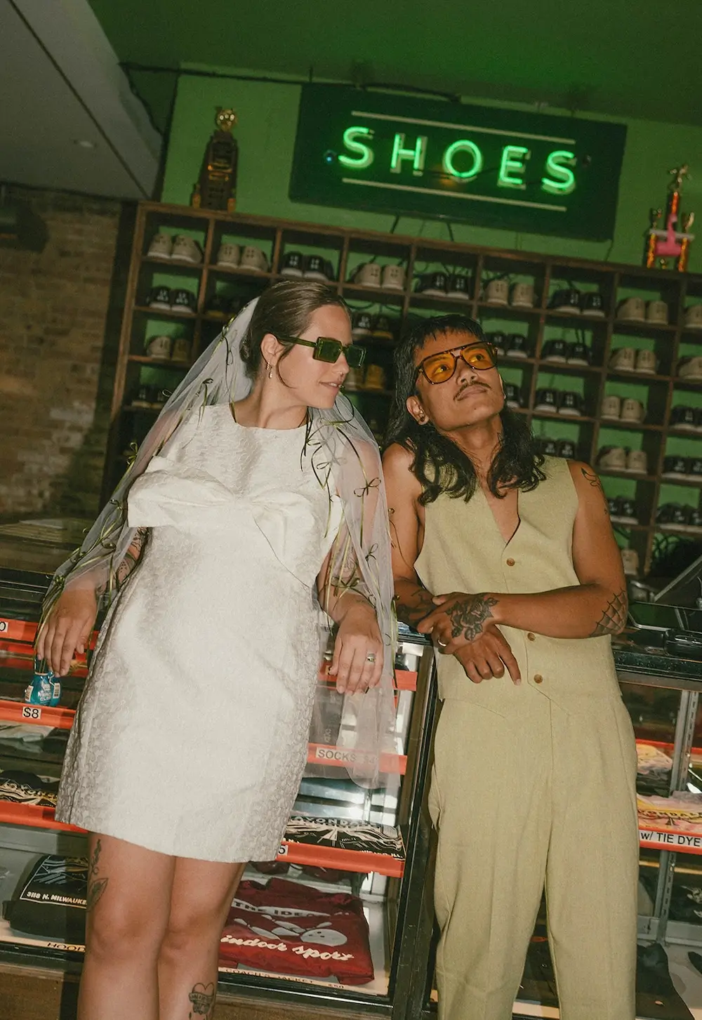 Couple in modern wedding attire and tinted sunglasses posing inside a vintage bowling alley beneath a neon “Shoes” sign with retro shoe racks in the background.