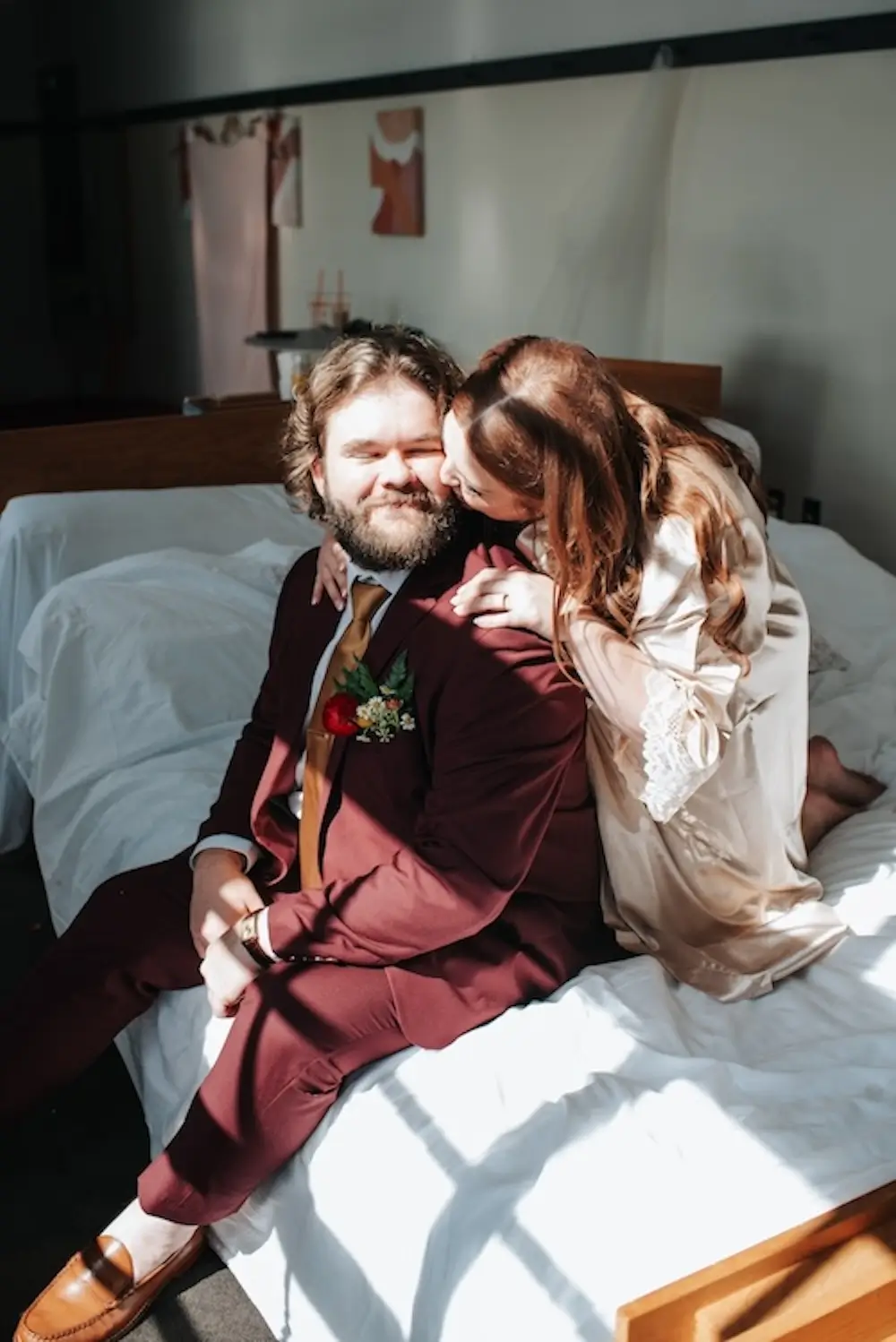 Bride in robe leaning over groom in burgundy suit as they sit on a bed during wedding day getting-ready moments.