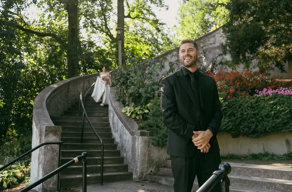 Groom waiting at the bottom of a curved stone staircase in a park as the bride walks down toward him for their first look.