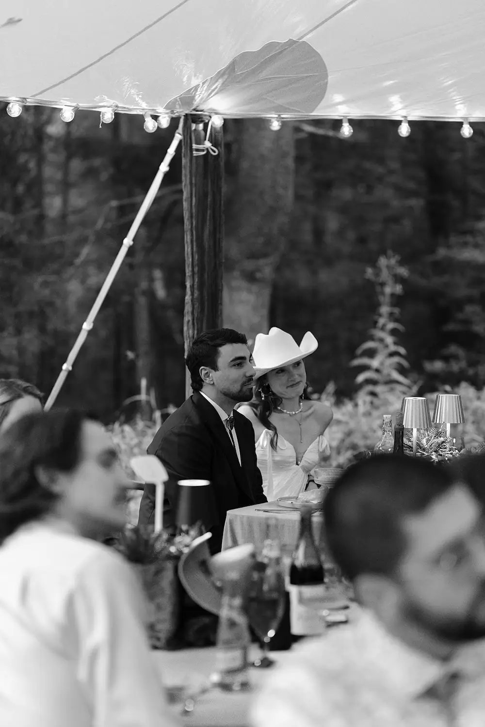 Bride in a cowboy hat and groom seated together at a candlelit outdoor reception dinner under a tent in the woods.