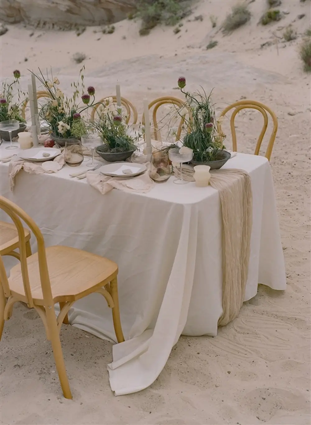 Beach wedding reception table set with natural linens, wild florals, and wooden chairs on sand.