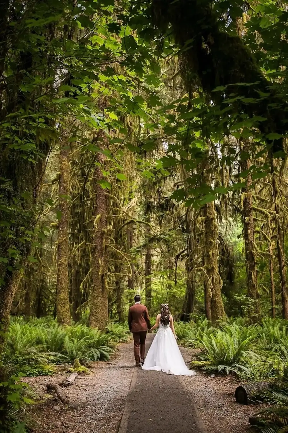 Bride and groom walking hand in hand along forest path surrounded by moss-covered trees.