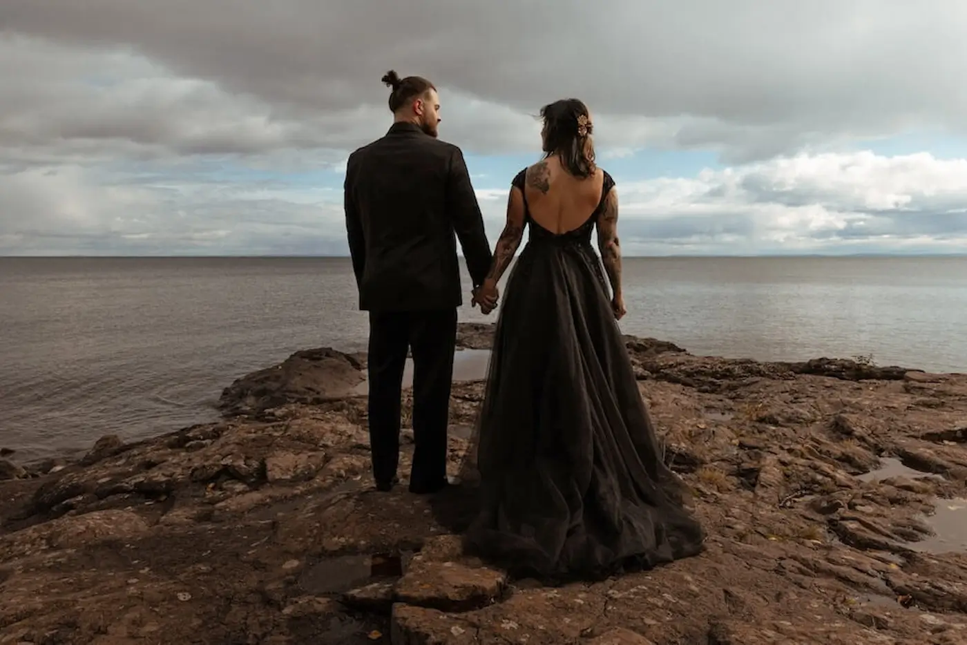 Couple in black wedding attire standing on rocky shoreline overlooking dramatic ocean landscape.
