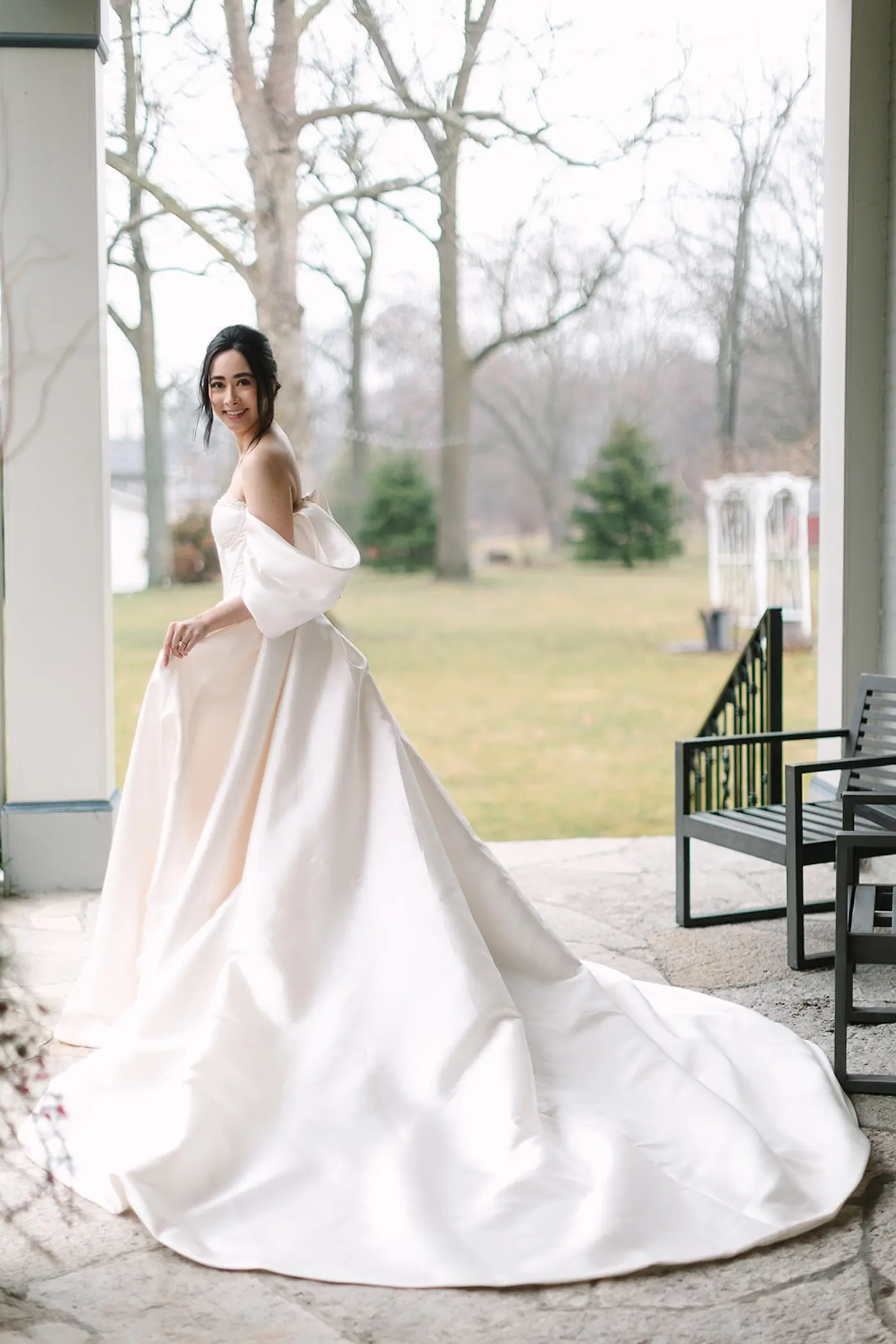 Bride in classic off-shoulder wedding gown standing on covered porch overlooking garden.
