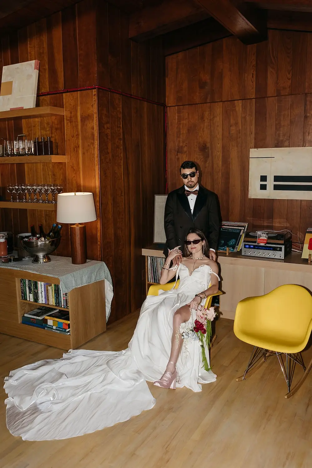 Bride in satin gown and groom in tuxedo posing in mid-century modern lounge with record player and wood paneling.