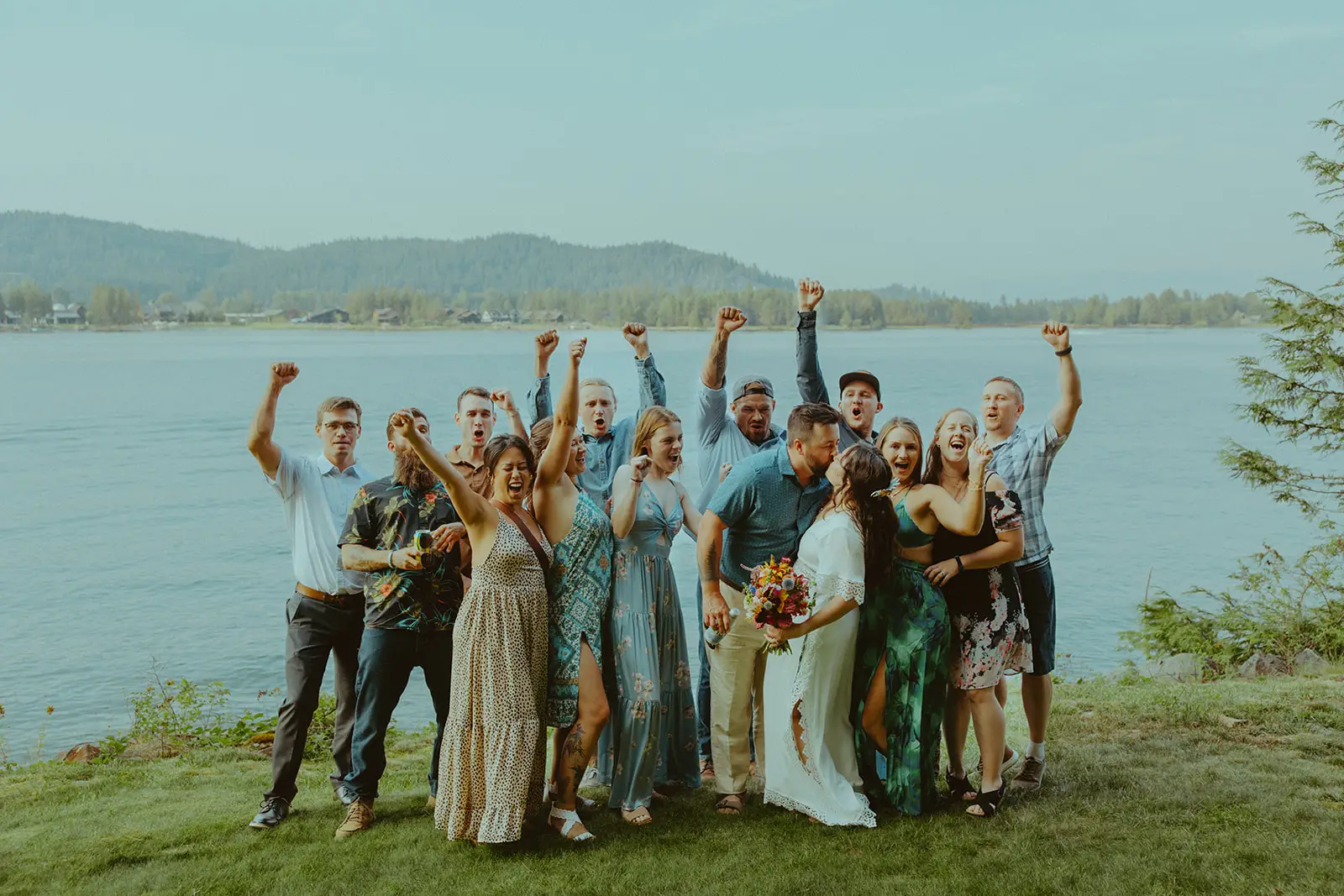 Group of wedding guests outdoors by a lake, cheering with raised arms, wearing a mix of summer dresses, short-sleeve shirts, and semi-casual attire on a grassy lawn