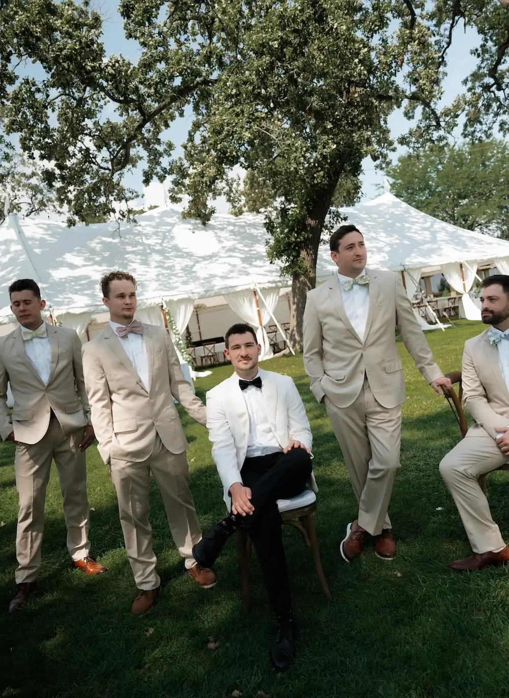 Group of groomsmen standing and sitting on a lawn under a tree, wearing light-colored suits with bow ties, with a white tent in the background