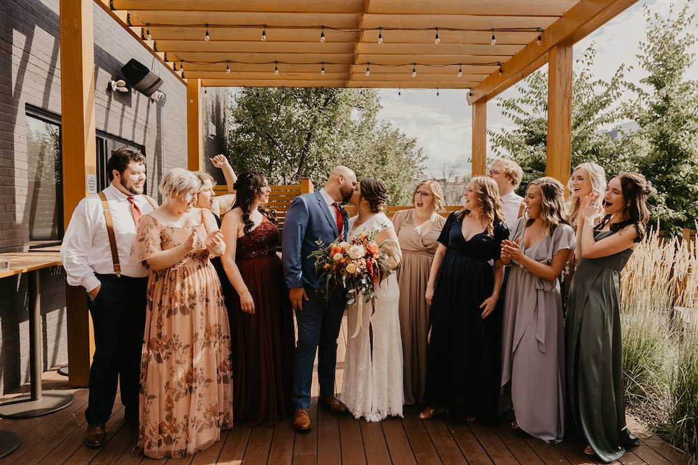 Wedding party gathered on a wooden patio under string lights, with guests wearing a mix of dresses and suits while the couple stands in the center holding a bouquet