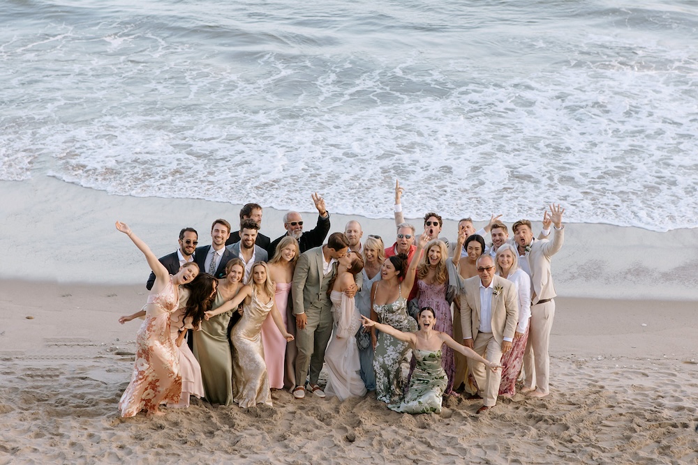 Group of wedding guests standing on a sandy beach by the ocean, wearing a mix of light-colored dresses, suits, and summer attire, with waves in the background