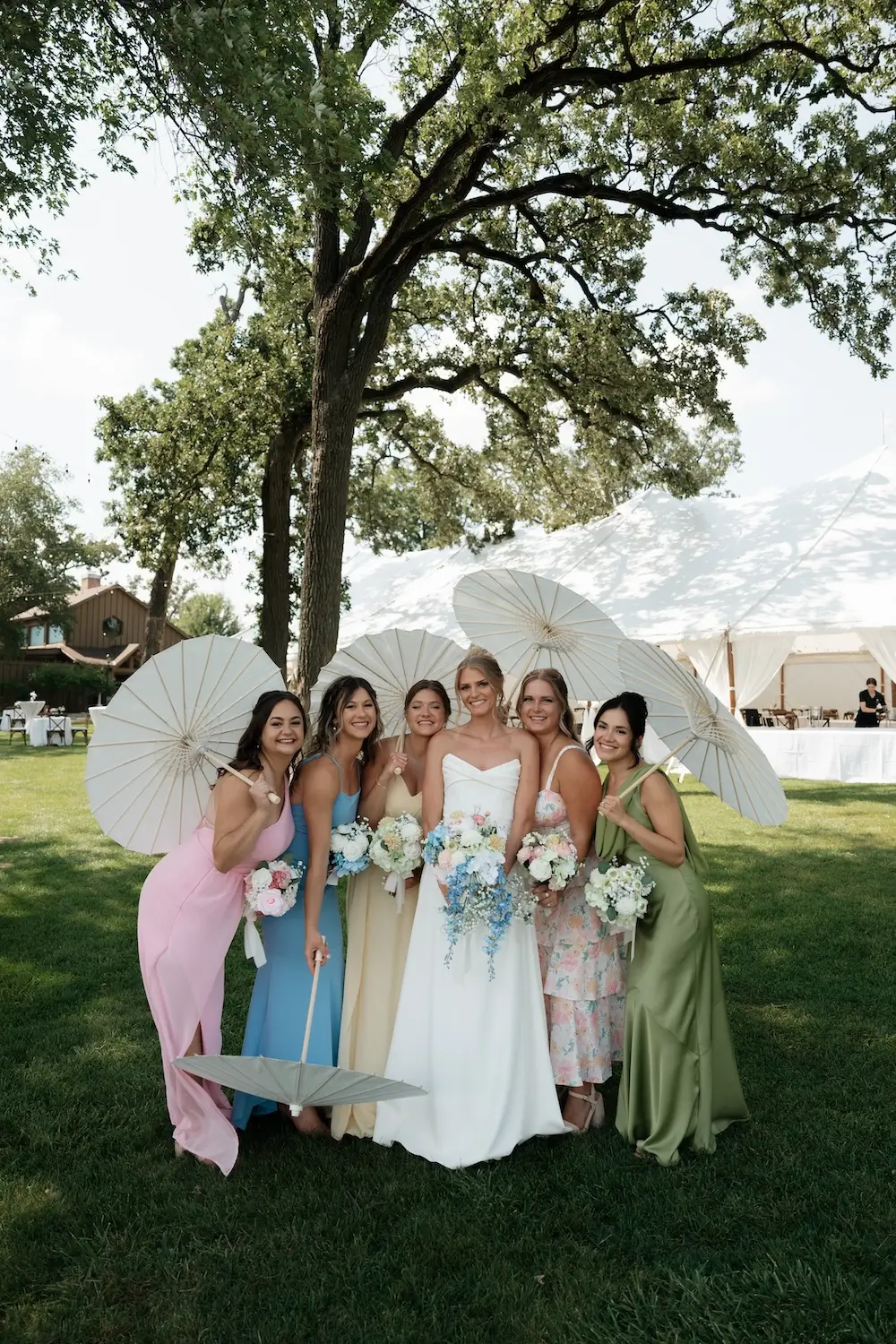 Bride and bridesmaids standing on a lawn under large trees, wearing pastel and light-colored dresses and holding parasols and bouquets in front of a white tent