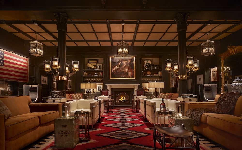 Wood-paneled wedding cocktail lounge with stone fireplace, leather sofas around a red geometric rug, American flag, and pendant candle lighting