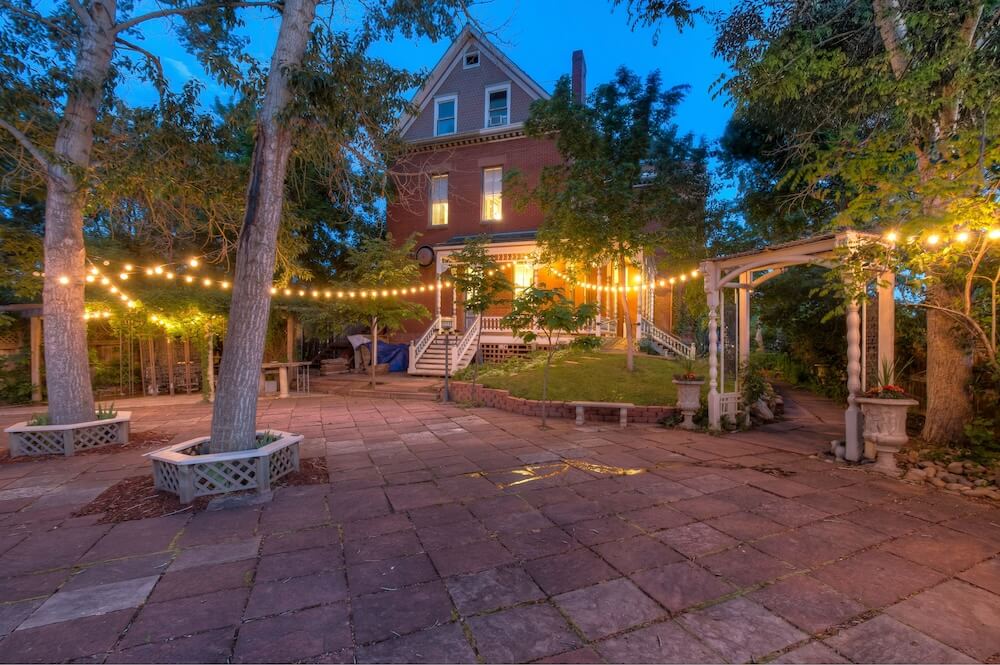 Three-story red brick Victorian small wedding venue at twilight, with a string-lit stone courtyard, white pergola, and shade trees