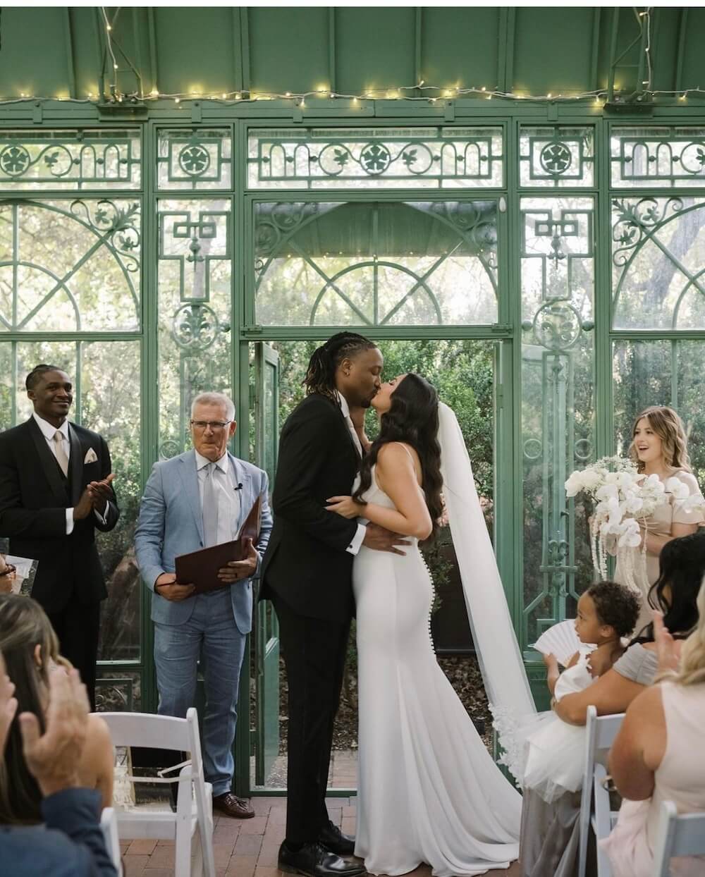 Micro wedding ceremony inside a green Victorian-style glass conservatory, couple kissing at the altar with overhead string lights and white florals