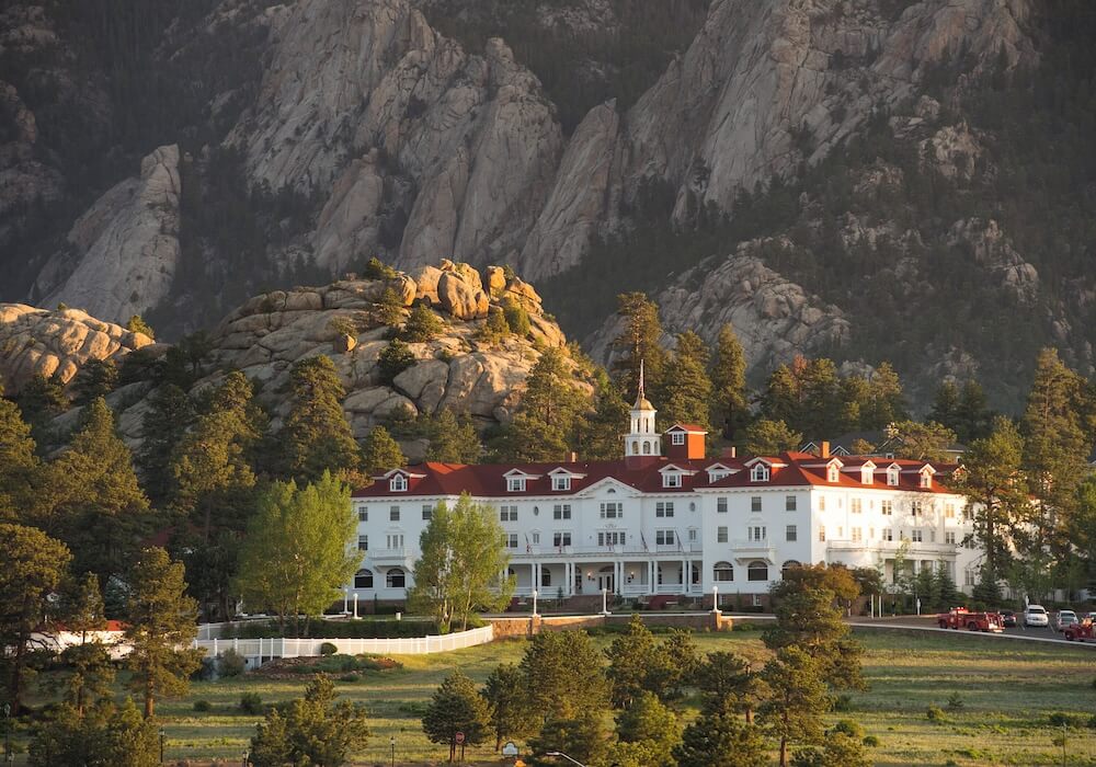 Large white wooden grand hotel wedding venue with red roof and central cupola, fronted by meadow and pine trees, rocky granite crags rising behind