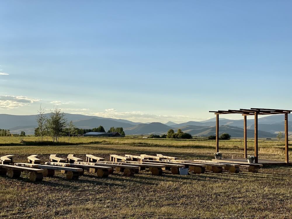 Outdoor ceremony space at a Colorado VRBO wedding venue, with a wooden arbor and rows of log benches facing distant hill ridges at sunset