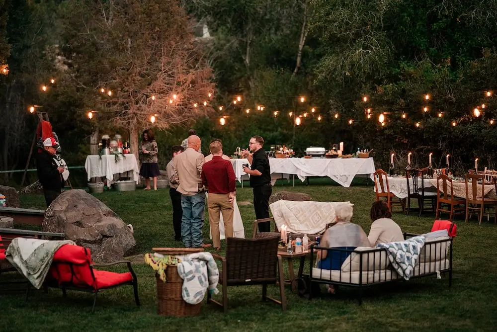 Outdoor evening micro wedding reception with overhead string lights, guests gathered around long candle-lit tables and lounge seating on the grass