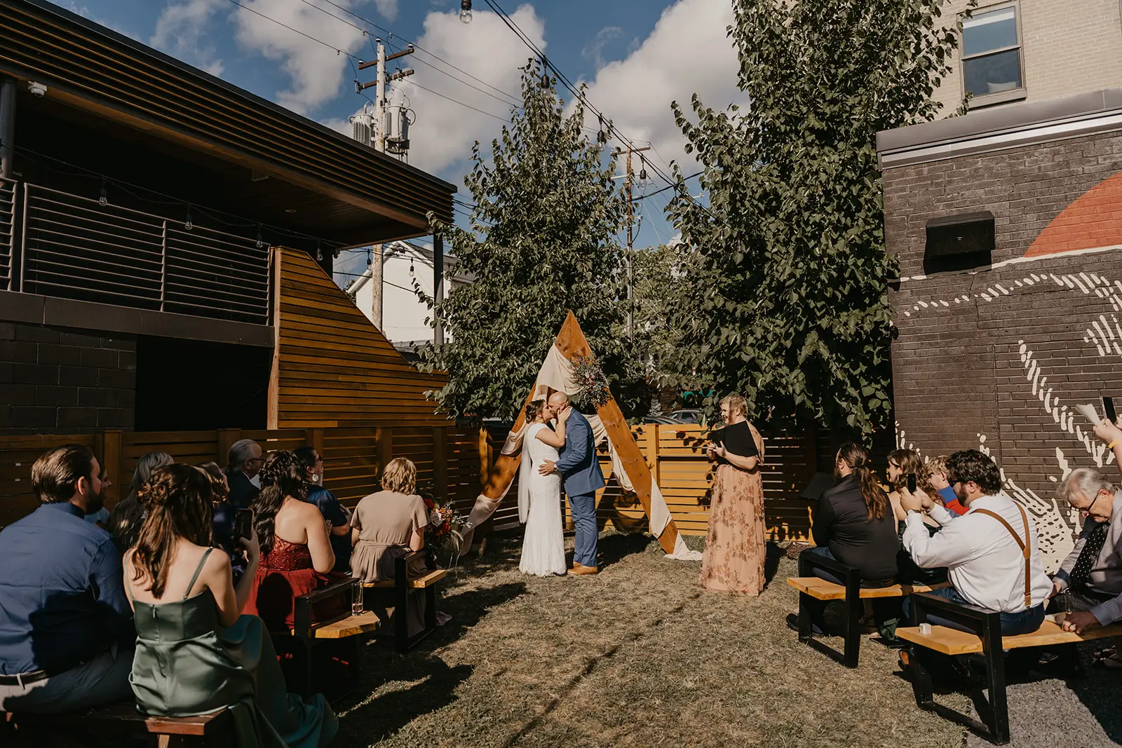 Urban backyard micro wedding ceremony with the couple kissing under a wooden A-frame arch, guests on benches and brick buildings on either side