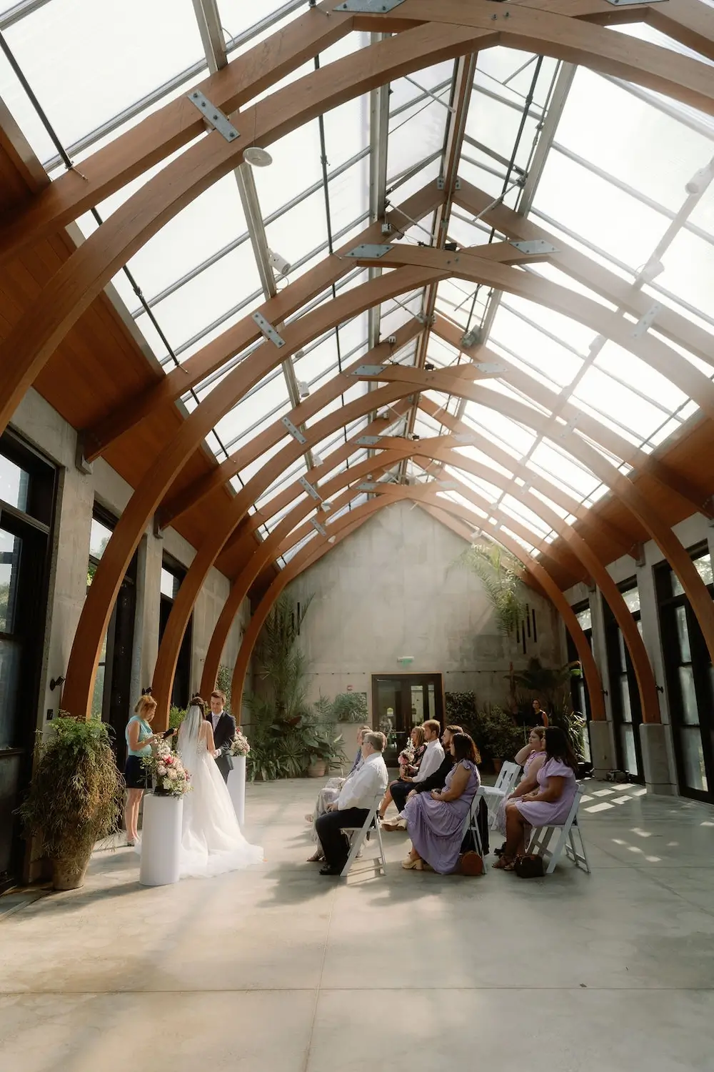 Intimate indoor micro wedding ceremony in a modern venue with a glass ceiling, wooden beams, and natural light.