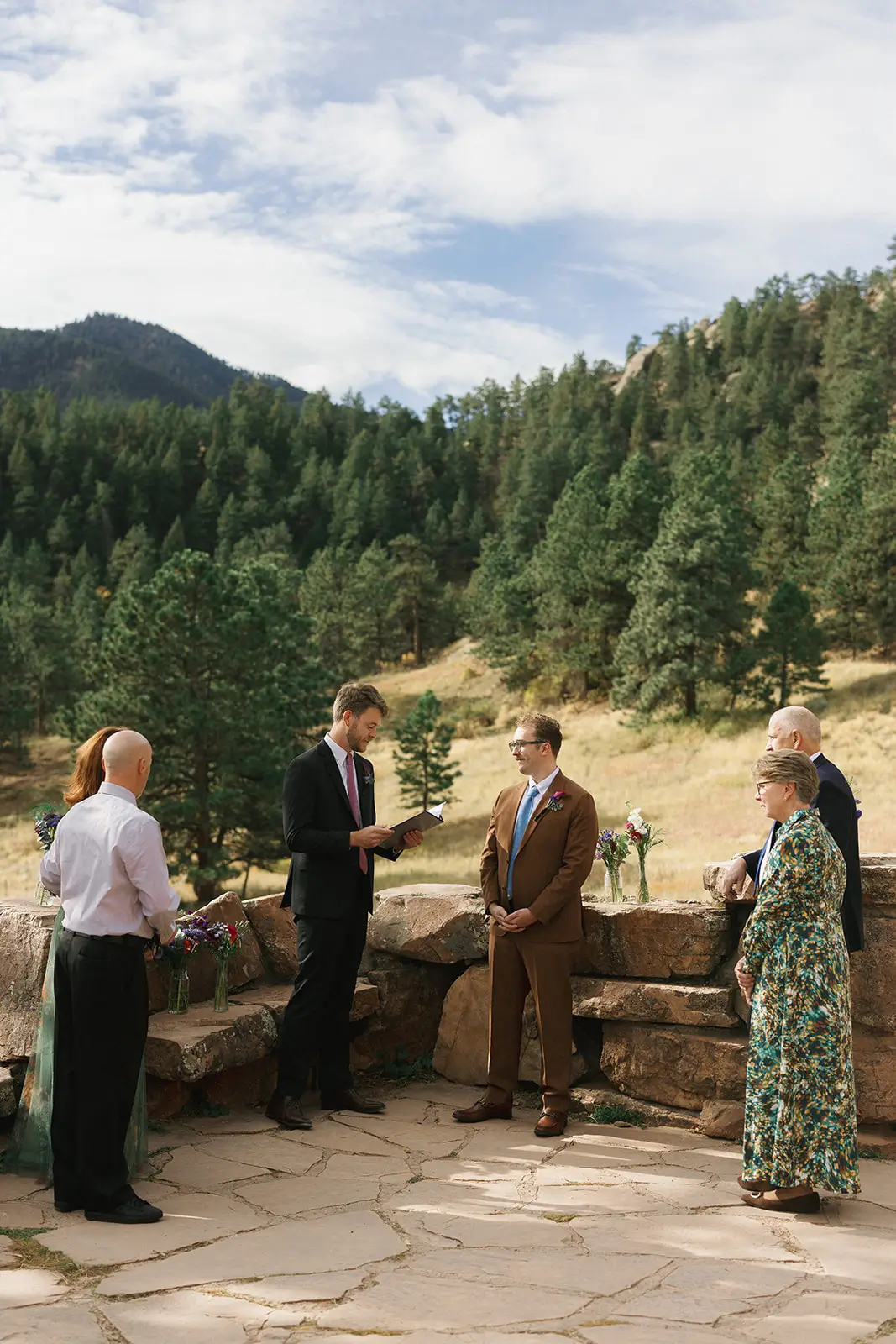 Outdoor micro wedding ceremony at a stone overlook patio, one partner reading vows from a folder, mountains and pine forest rolling away behind