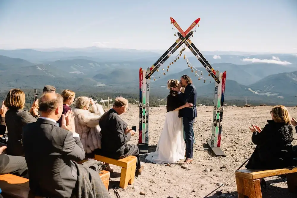 Mountain-top micro wedding ceremony with couple kissing under an arch of crossed skis, guests applauding on log benches and panoramic ridges behind