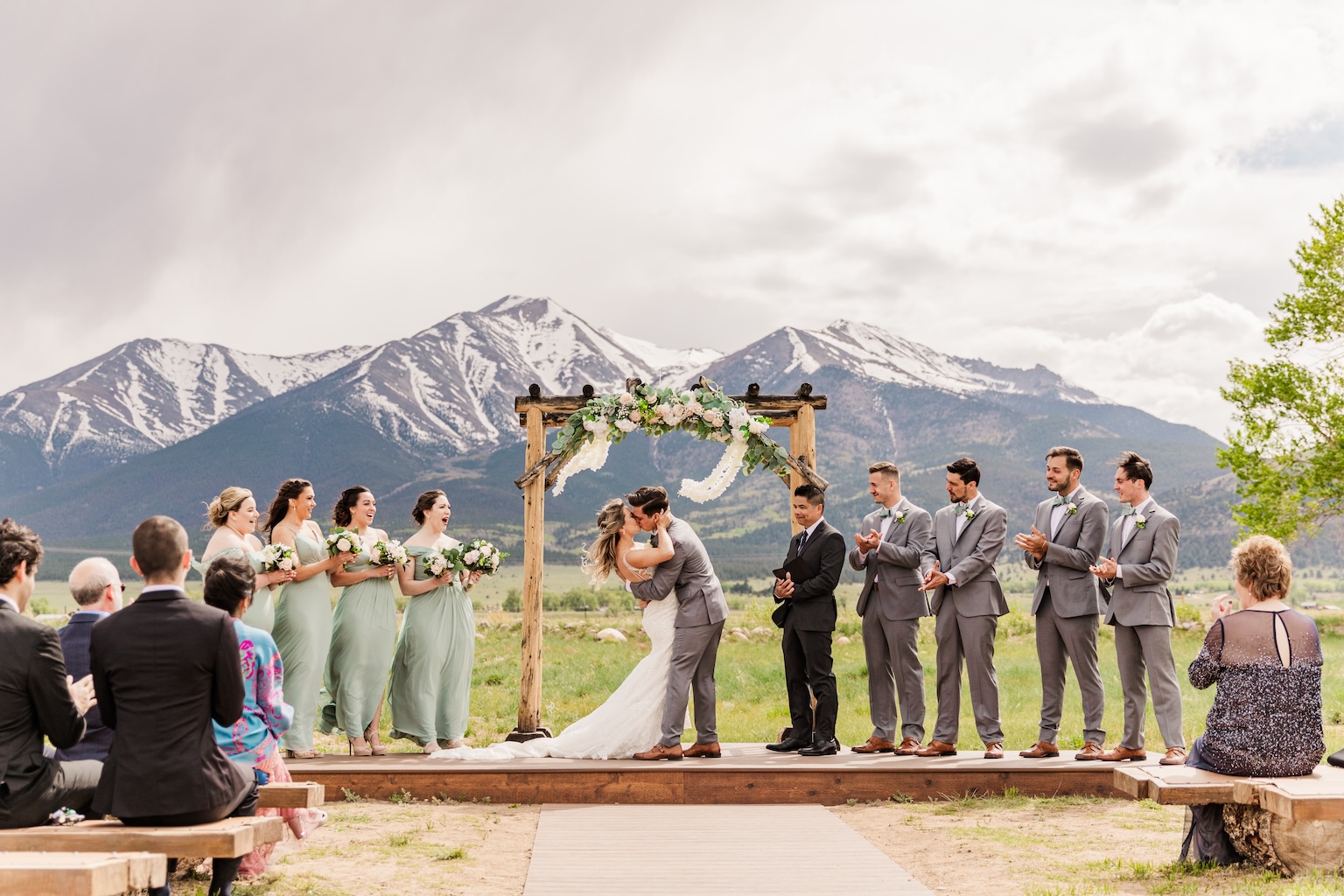 Micro wedding ceremony kiss under a wooden floral arch, snow-capped Colorado mountains behind, bridesmaids in sage and groomsmen in gray clapping