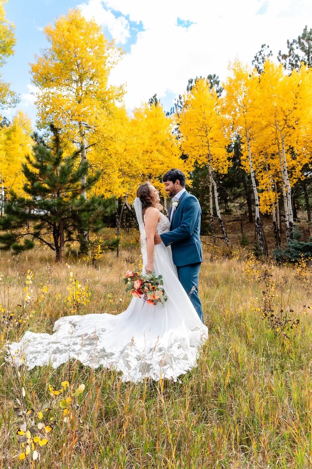 Couple facing each other in a meadow ringed by golden aspens, bride in a long-train lace gown holding a peach bouquet, groom in a blue suit