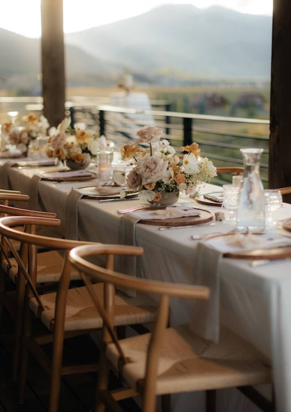 Micro wedding reception table on a covered mountain-view deck, wishbone chairs, blush and cream florals and votive candles in golden-hour light