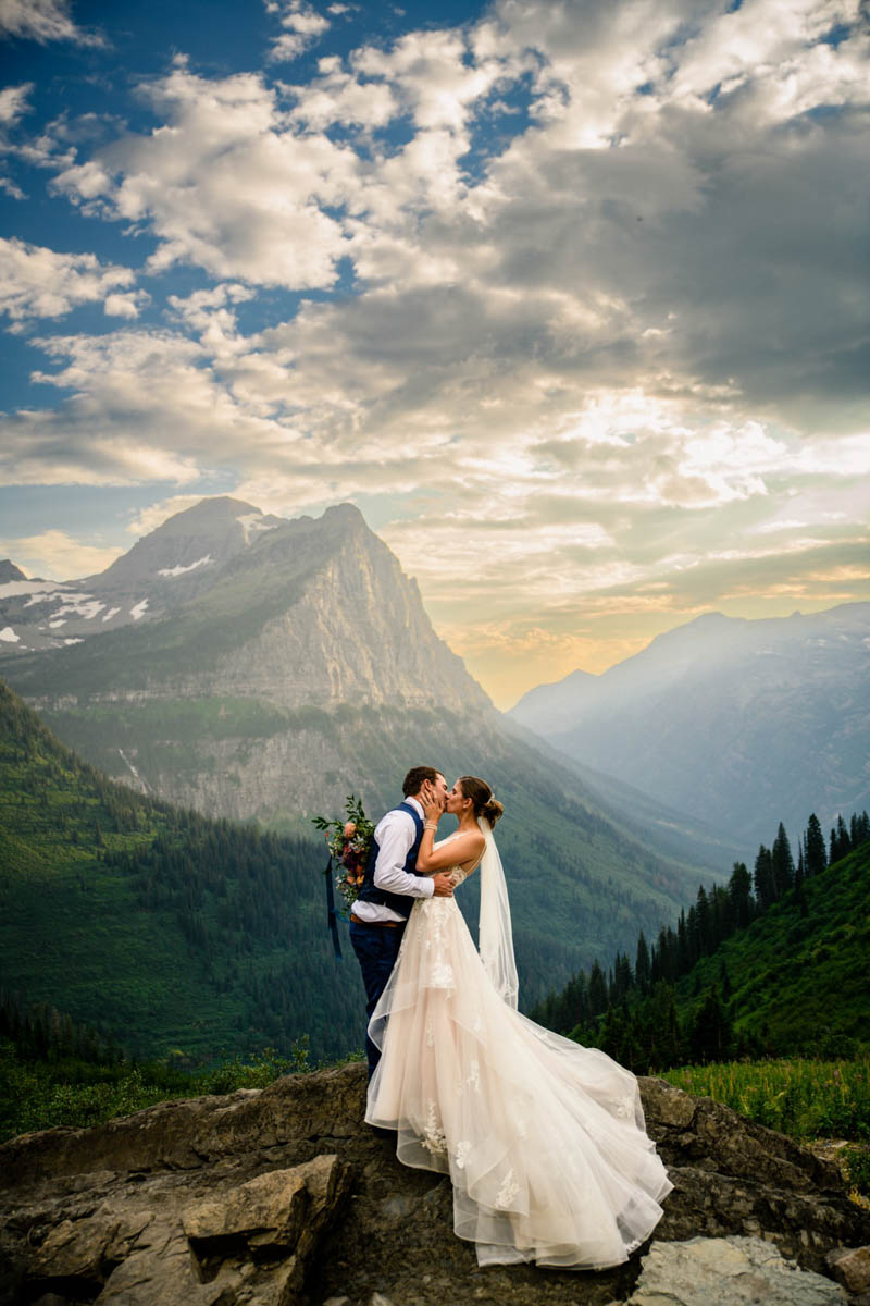 Elopement couple kissing on a rocky cliff above a forested valley in Glacier National Park, dramatic granite peak and sunbeam-streaked clouds behind