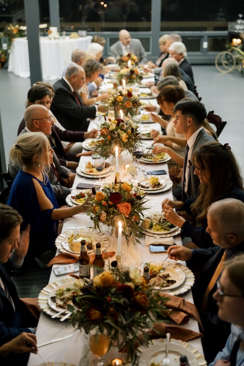 Long candlelit wedding reception table with about twenty guests dining, autumn floral runners in orange and burgundy with copper-toned napkins