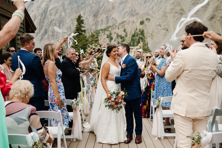 Mountain ceremony recessional on a wooden deck, couple walking back up the aisle as guests wave white ribbon wands, sheer granite peak rising behind