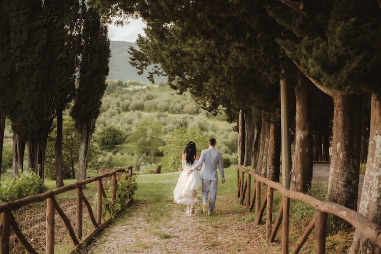 Couple walking down a cypress-lined Tuscan path during an Italian destination wedding, rolling green hills beyond a wooden post-and-rail fence