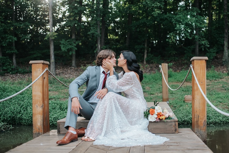 Couple kissing on a wooden dock with rope railings beside a forest pond, bride in long-sleeve sequin gown with peach bouquet, groom in gray suit