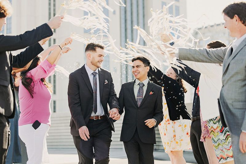 Courthouse micro wedding exit with a streamer toss, both grooms in dark suits hand-in-hand as guests cheer with white paper streamers