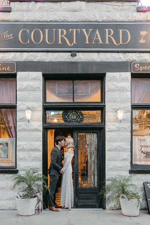 Couple kissing in the doorway of a historic stone-front small wedding venue with gold-lettered signage and basket planters flanking the entry
