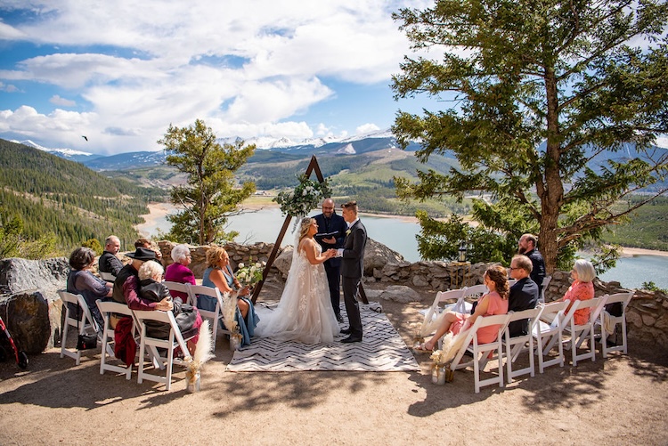 Outdoor small wedding ceremony at a wooden triangle altar on a mountain overlook, with a dozen guests seated above an alpine lake