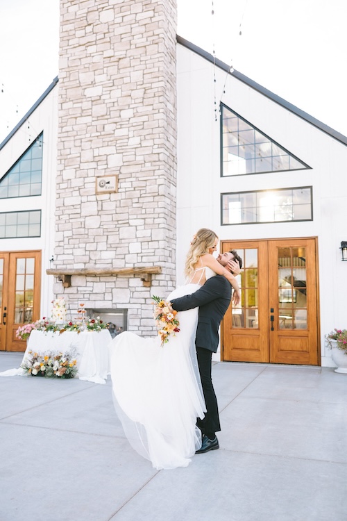 Couple on the patio of a white board-and-batten wedding venue with a tall stone chimney and wooden double doors, groom lifting bride mid-twirl