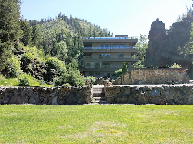 Multi-story canyon lodge wedding venue between a pine hillside and rocky outcropping, with a stone-walled terrace and wide grass lawn in summer light