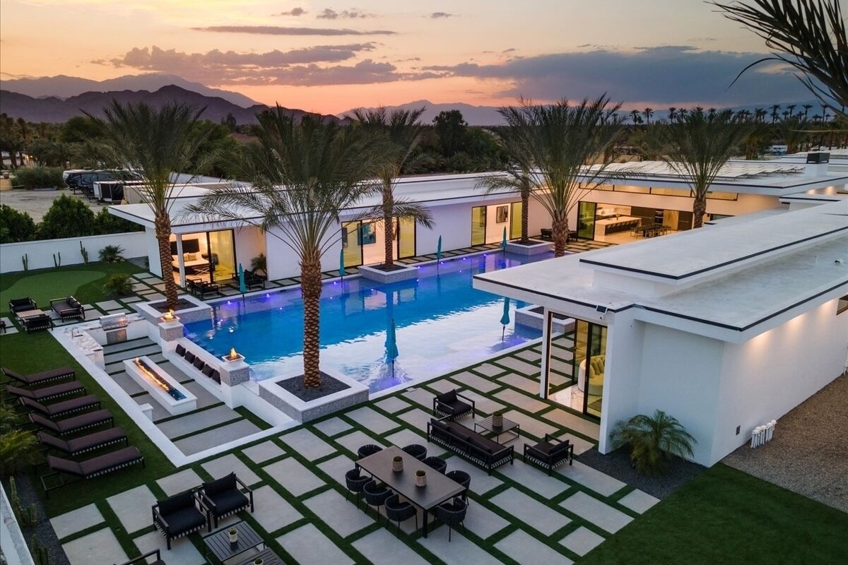 Modern desert VRBO wedding villa at twilight with a blue-lit pool, palm-island patio, linear fire feature, and multiple dining and lounge zones
