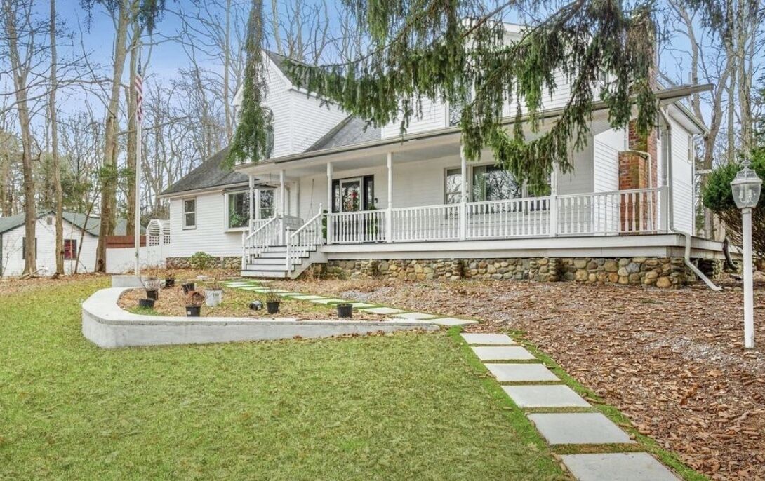 White clapboard lakehouse wedding venue with a wide covered porch, fieldstone foundation, brick chimney, and stepping-stone path through a wooded yard