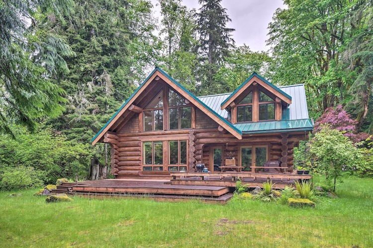 Two-story log cabin wedding venue with teal metal roof, floor-to-ceiling windows, and a wraparound deck surrounded by Pacific Northwest evergreens