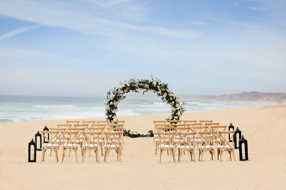 Beachfront ceremony with a circular floral arch on pale sand, rows of wood crossback chairs flanked by black lanterns, and surf and cliffs behind