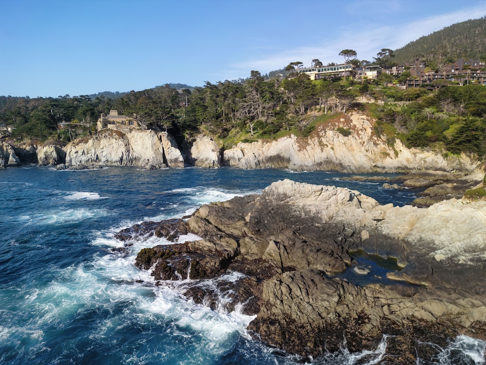 Aerial Carmel coastline with jagged sea cliffs and crashing surf, the cliff-top resort tucked among cypress on a wooded bluff in the distance