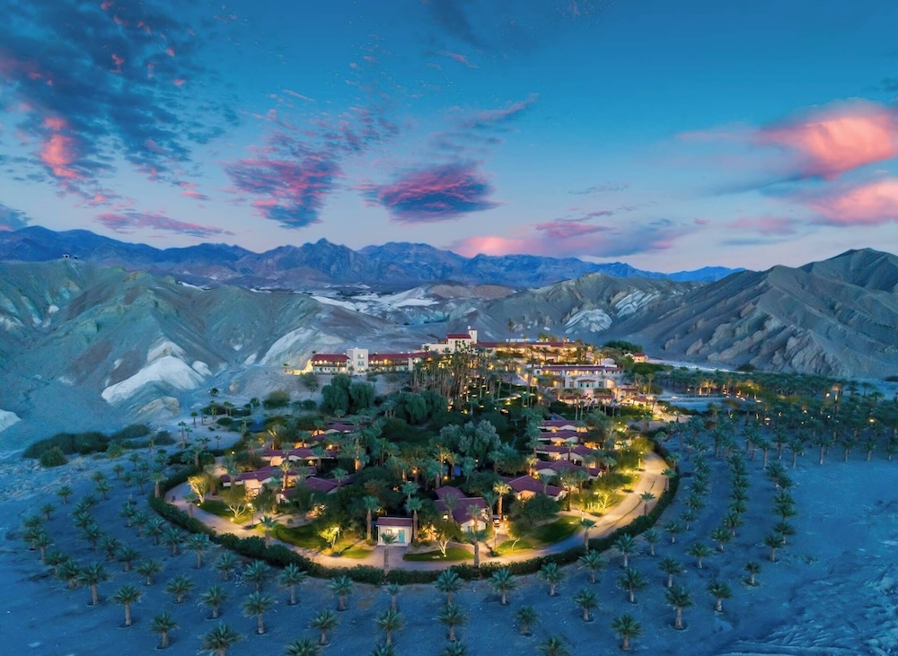 Aerial dusk over a desert oasis resort ringed by date palms, mission-style inn glowing among adobe casitas, eroded hills and pink sunset behind
